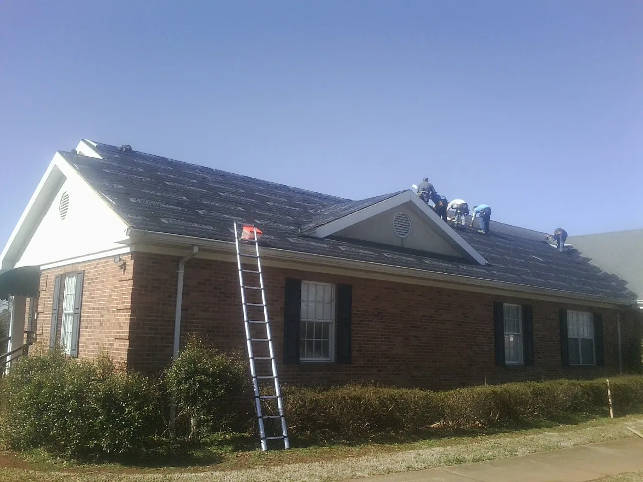 Crew tearing shingles off a brick ranch roof with a ladder set — Emergency Roof Repair work by our crew in Windsor Heights