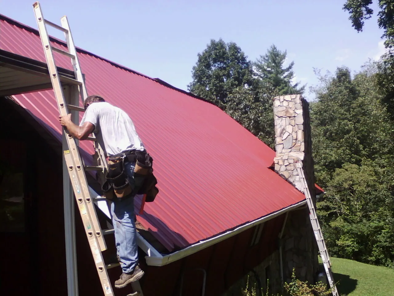 Roofer on a ladder beside a red metal roof and stone chimney — Soffit & Fascia Repair by Roofing Brothers in Windsor Heights